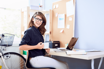 Smiling woman with glasses wearing blue shirt and white pants sitting at a desk with laptop open holding a cup of coffee.